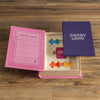 The Board Game: Candy Land Vintage Bookshelf Edition is open on a wooden surface, featuring colorful pieces and cards arranged in a pink and purple book-style box—a perfect family-friendly preschool game.