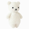 The Cuddle + Kind Baby Bouclé Bear - Ivory is a plush white teddy bear with black eyes, a small black nose and mouth, and pink blush spots on its cheeks, hand-knit from Peruvian alpaca yarn, standing upright against a white background.