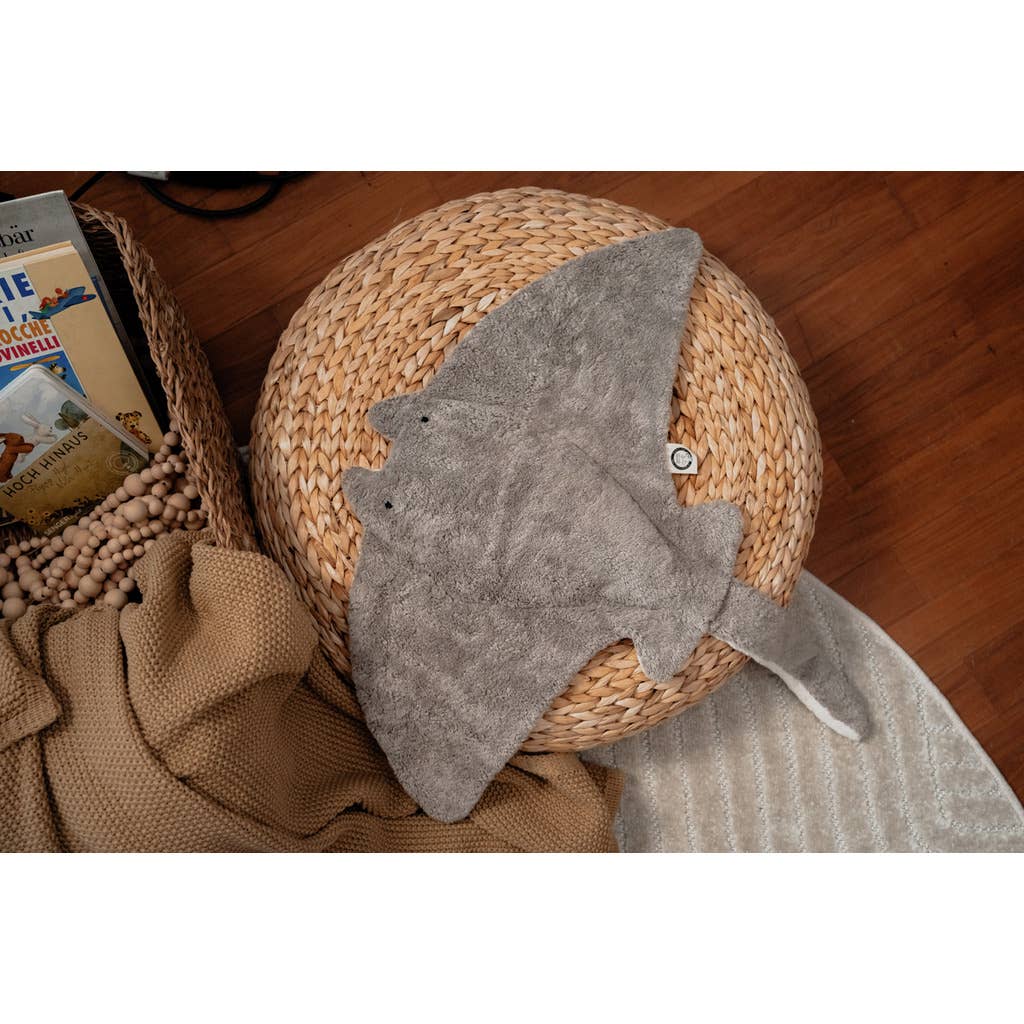 The Senger Naturwelt Stuffed Animal - Large Manta Ray sits atop a round woven ottoman beside a basket of books, a beaded garland, and a beige blanket on wooden floors—an ideal accent for cozy, sustainably styled spaces.