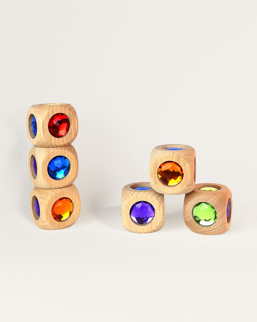 Two stacks of six light-colored Bauspiel Magic Cube, 1-Piece wooden cubes on a light surface, each featuring a large, faceted, colorful jewel embedded in the center.