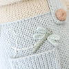 Close-up of light blue knit fabric with a small embroidered dragonfly and white stitched line; a beige button and cream knitting from the fair trade, hand-knit Cuddle + Kind Bailey The Mouse doll appear in the background.
