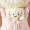 Cuddle + Kind Briar The Bunny - Peony Pink is a small, cream-colored hand-knit bunny with floppy ears and a smiling face, sitting in the front pocket of a pink knit garment, highlighting sustainable fair trade craftsmanship.