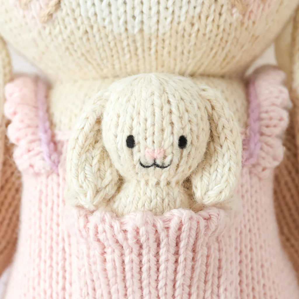 Cuddle + Kind Briar The Bunny - Peony Pink is a small, cream-colored hand-knit bunny with floppy ears and a smiling face, sitting in the front pocket of a pink knit garment, highlighting sustainable fair trade craftsmanship.