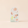 The illustrated Cuddle + Kind Tiny Flora the Duckling (Mauve) book showcases a floral cover with a small figure, revealing whimsical artwork of cartoonish ducks, landscapes, and imaginative scenes. Its pastel hues and elegant design capture the charm of handmade dolls.