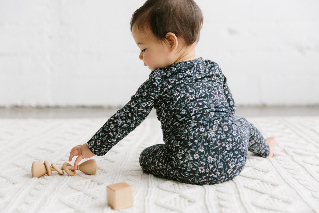 A baby in a floral jumpsuit plays with a Wooden Baby Rattle on a white textured rug, sitting with their back to the camera.