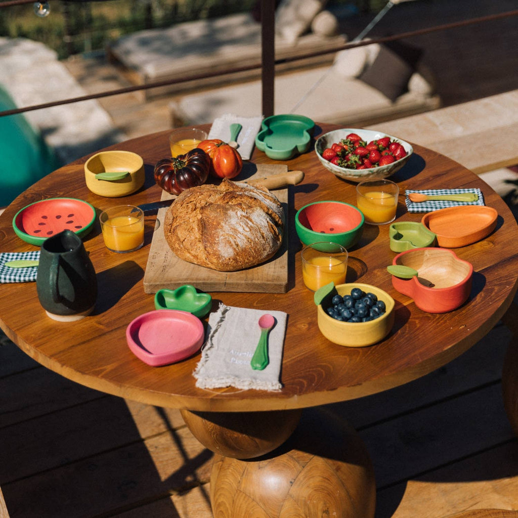 A round wooden table set for breakfast features a loaf of bread, fruit bowls, orange juice, colorful plates and cups, utensils, and the Wally the Watermelon Bowl & Spoon Set with strawberries—all beautifully arranged outdoors on a sunny day.