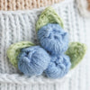Close-up of three blue yarn balls styled as blueberries with two green leaves, hand knit from cotton and attached to a white fabric background, featured alongside Cuddle + Kind Asher The Cow.
