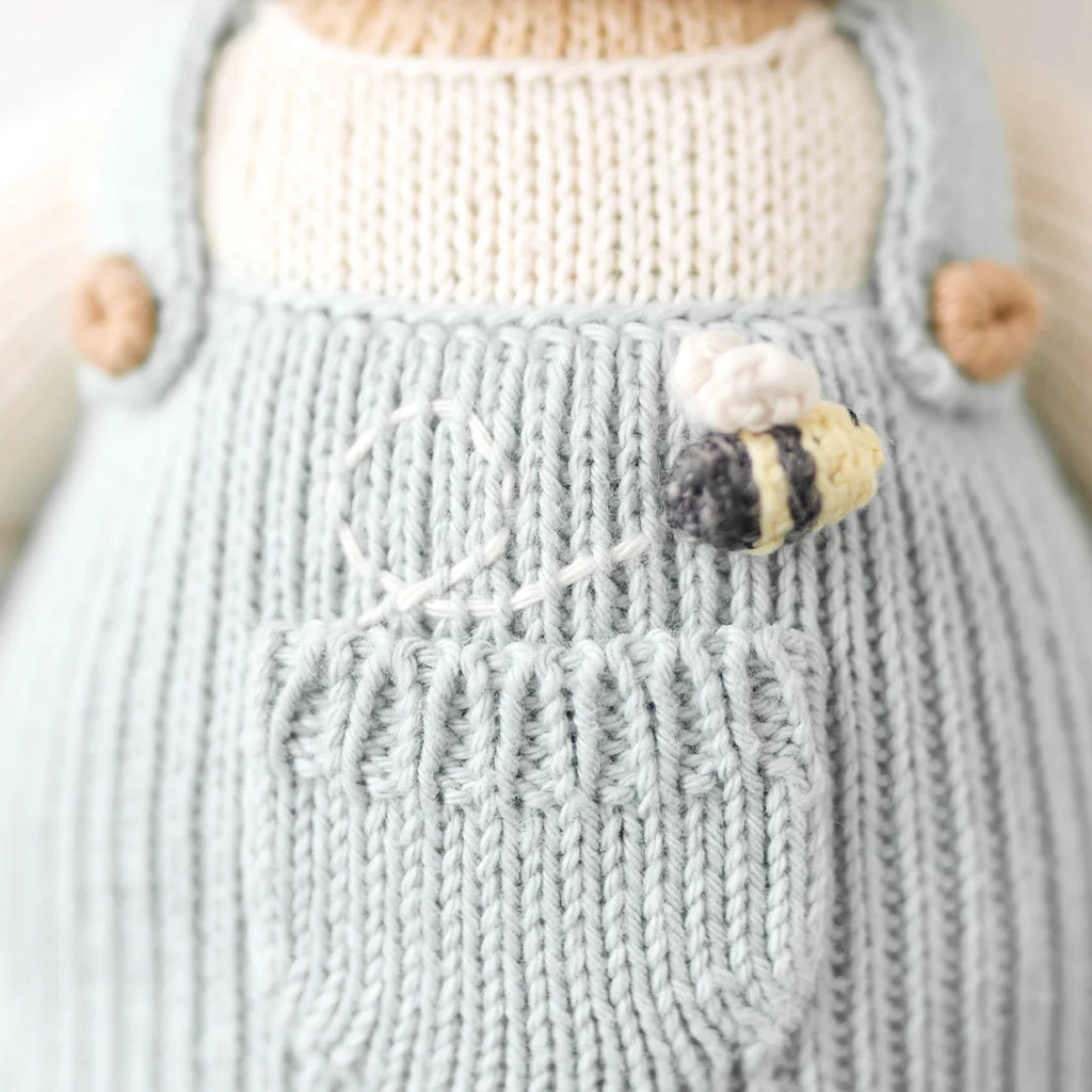 Close-up of a hand-knit Cuddle + Kind Charlie The Honey Bear outfit in light blue with a front pocket, featuring an embroidered bee and a white stitched design above the pocket.
