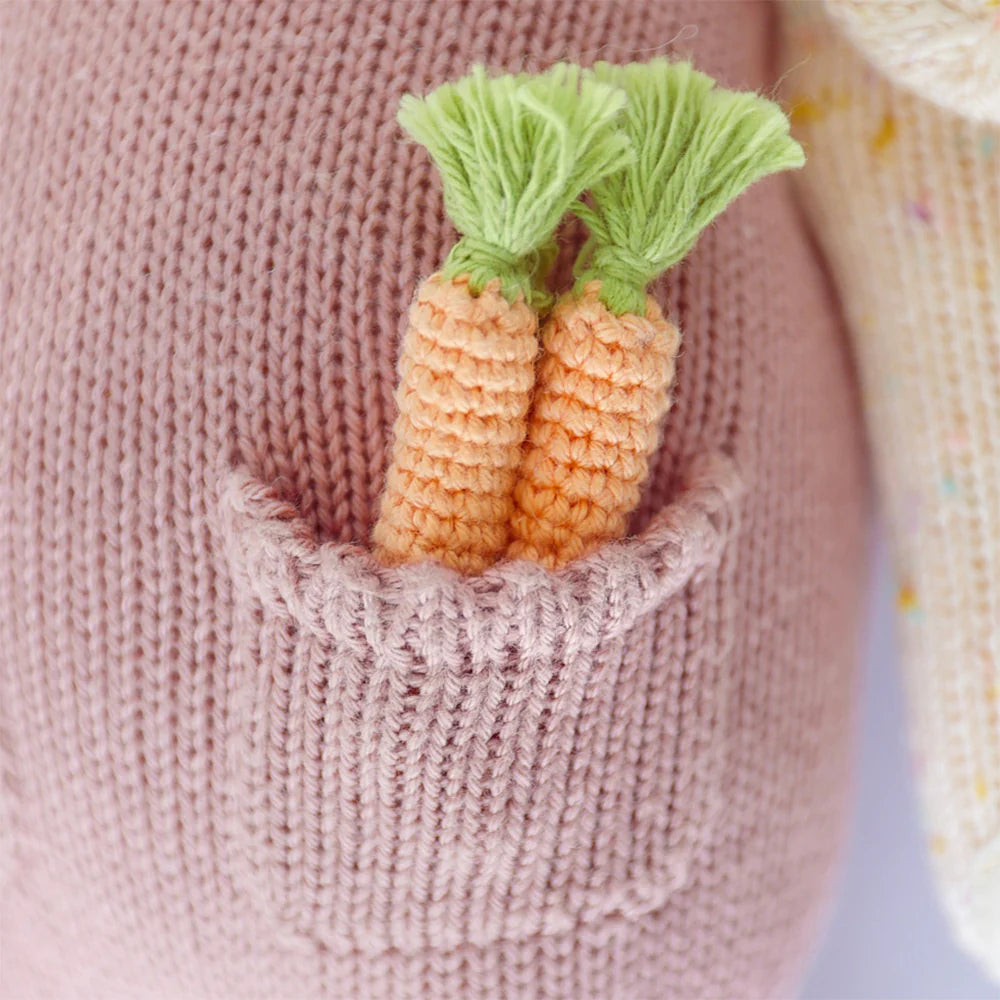 Two small, hand-knit carrots with green tops are tucked into a pink cotton pocket on Cuddle + Kind Harper the Bunny.