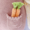 Two small, hand-knit carrots with green tops are tucked into a pink cotton pocket on Cuddle + Kind Harper the Bunny.
