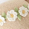 Close-up of Cuddle + Kind Olivia The Honey Bear’s beige hand-knit fabric, decorated with white flowers with yellow centers and green leaves arranged in a row.