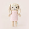 A Cuddle + Kind Tiny Hannah The Bunny hand-knit, cream-colored bunny doll wearing a light pink dress. The non-toxic bunny has embroidered facial features and a garland of white flowers stitched on its head. It stands upright against a plain, light background, embodying fair trade practices.