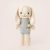 A Cuddle + Kind Tiny Henry The Bunny from the cuddle+kind dolls collection, wearing a light blue romper with a small embroidered carrot on the front. The safety certified bunny has long, floppy ears, a stitched smile, and black eyes against a plain, light-colored background.