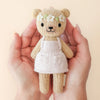 Hands cradle a charming, hypoallergenic Cuddle + Kind Tiny Olivia the Honey Bear. With a beige body, white apron, and flower headband, the bear's warm smile shines against a soft peach background, epitomizing cozy fair trade craftsmanship.