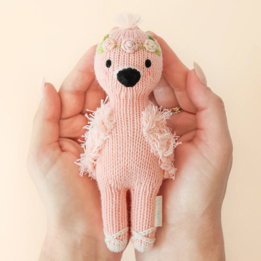 Hands gently holding the Cuddle + Kind Tiny Penelope the Flamingo, a small, safety-certified knitted pink flamingo toy with a floral headband, handmade in Peru.