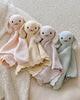 Four Cuddle + Kind Organic Cotton Baby Bunny Loveys (Blush), featuring embroidered faces and floppy ears, are laid side by side on a textured, cream-colored surface.