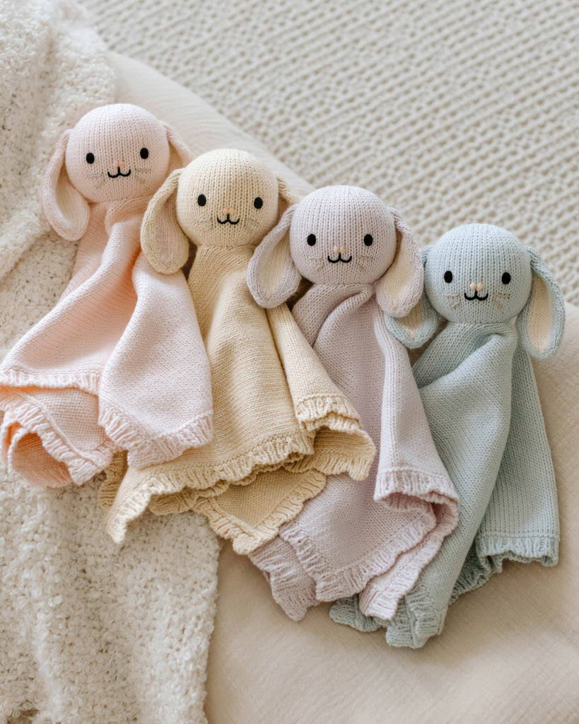 Four Cuddle + Kind Organic Cotton Baby Bunny Loveys (Blush), featuring embroidered faces and floppy ears, are laid side by side on a textured, cream-colored surface.