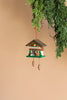 A Christian Ulbricht Ornament – Cuckoo with Deer, crafted in Germany, hangs from a branch against a light beige background. This decorative piece features a quaint wooden house design adorned with pine trees, a deer, and two pinecone weights. It is important to note that this charming item is not a toy.
