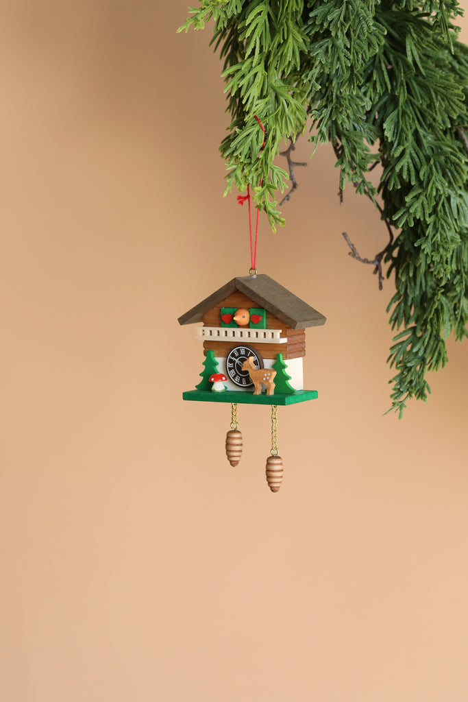A Christian Ulbricht Ornament – Cuckoo with Deer, crafted in Germany, hangs from a branch against a light beige background. This decorative piece features a quaint wooden house design adorned with pine trees, a deer, and two pinecone weights. It is important to note that this charming item is not a toy.