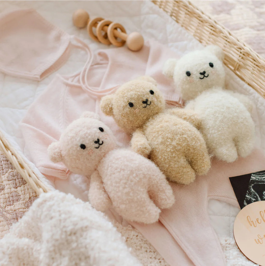 A Cuddle + Kind Baby Bouclé Bear - Honey sits in a wicker basket with soft baby clothes, a wooden rattle, and a quilted blanket, creating a cozy and charming nursery scene.