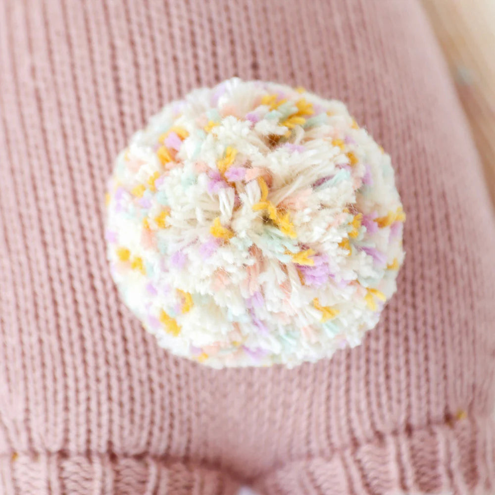 A multicolored, fluffy yarn pom-pom tops a pink knitted surface, bringing a playful accent to Cuddle + Kind Harper the Bunny, a premium cotton doll handmade in Peru.