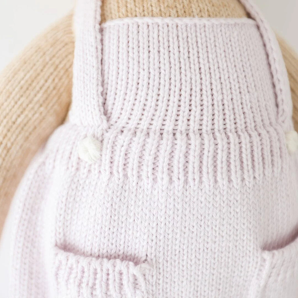 Close-up of a hand-knit light pink garment with two small pockets and straps, displayed on a beige textured surface, worn by Cuddle + Kind Olivia The Honey Bear doll.
