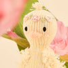 The Cuddle + Kind Tiny Flora the Duckling (Mauve) is a knitted duck with a fluffy texture, adorned with floral head decorations, surrounded by pink flowers and green leaves against a light background, showcasing sustainable toy craftsmanship.