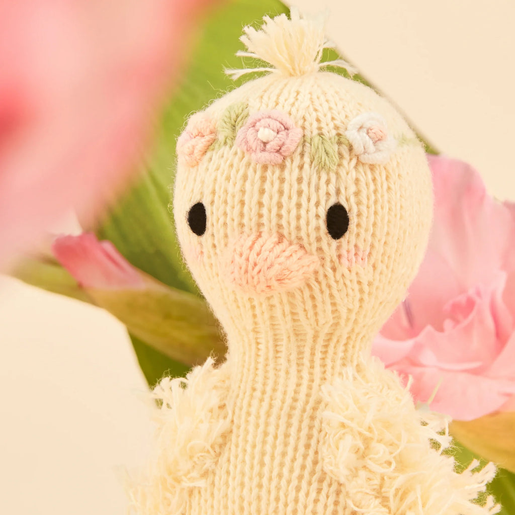 The Cuddle + Kind Tiny Flora the Duckling (Mauve) is a knitted duck with a fluffy texture, adorned with floral head decorations, surrounded by pink flowers and green leaves against a light background, showcasing sustainable toy craftsmanship.