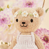 Tiny Olivia the Honey Bear by Cuddle + Kind, a hypoallergenic knitted teddy with a floral headband and white apron, stands smiling with black stitched eyes amid pink and white dahlias against a neutral background, offering gentle playtime fun.