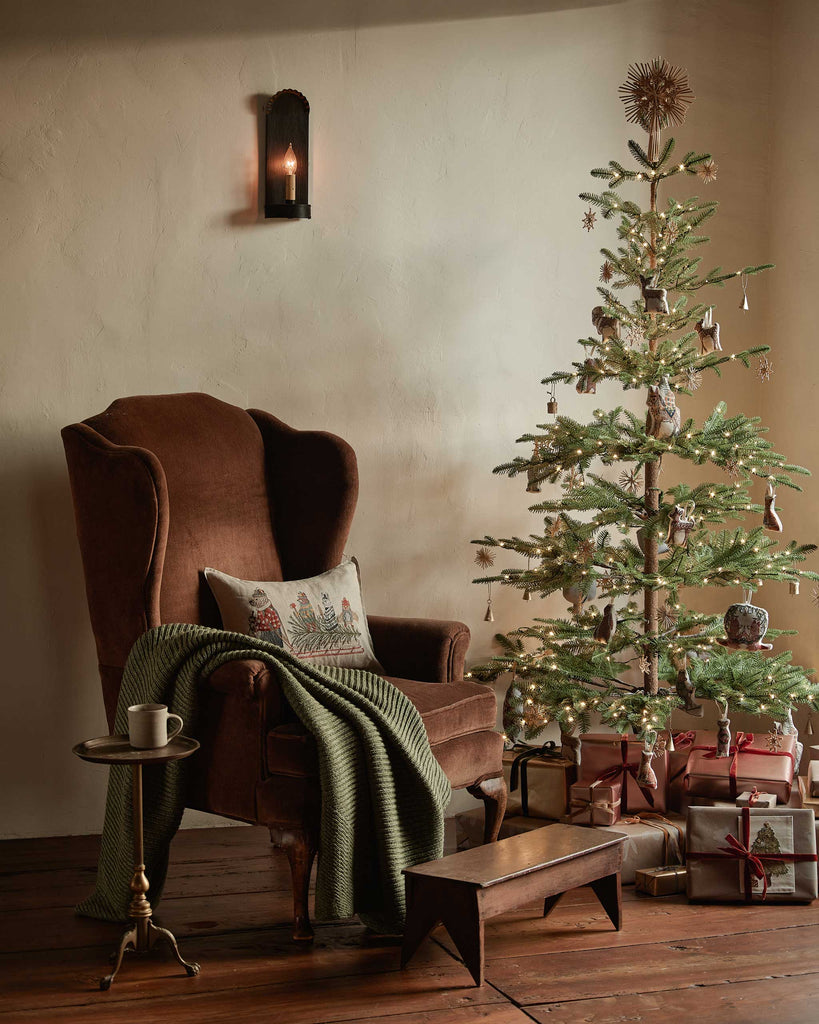 Coral & Tusk Christmas Tree Sled Pillow featuring embroidered animals on a rectangular decorative pillow, resting on a brown wingback chair next to a decorated Christmas tree.