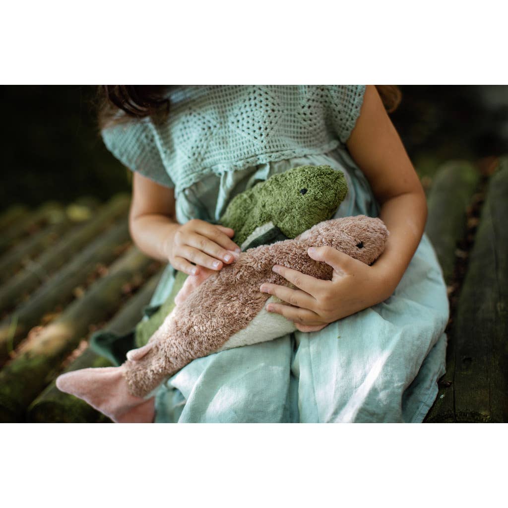 A child in a light blue dress sits outdoors, holding two Senger Naturwelt Cuddly Animal - Fish plush toys—one green and one pink—on their lap.