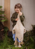 A young child in a green sweater and blue pants smiles while holding a large Senger Naturwelt Cuddly Animal - Linen Goose upright by its neck, standing outside on grass near a wall and wooden door.