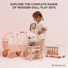 A young girl holds a cloth doll next to a wooden doll pram, a high chair, and a Wooden Dolls Cot with soft bedding. Text reads “Explore the complete range of wooden doll play sets” and “All sold separately.”