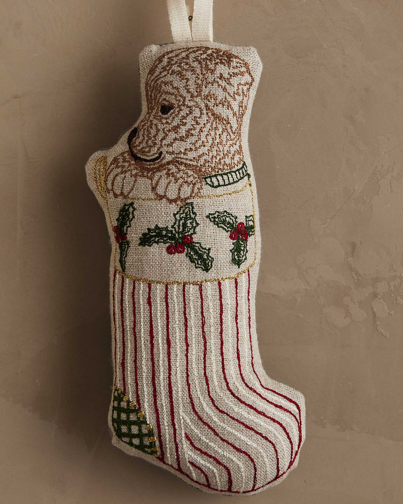 The Coral & Tusk Puppy Stocking Ornament features an embroidered puppy with holly leaves and red stripes, bringing a cheerful holiday touch to your décor when hung against a beige wall.