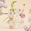 A Cuddle + Kind Tiny Olivia the Honey Bear, in a white dress and hypoallergenic design, sits next to a floral decorative box. In the background, a bouquet of pink and white flowers enhances the light beige setting.