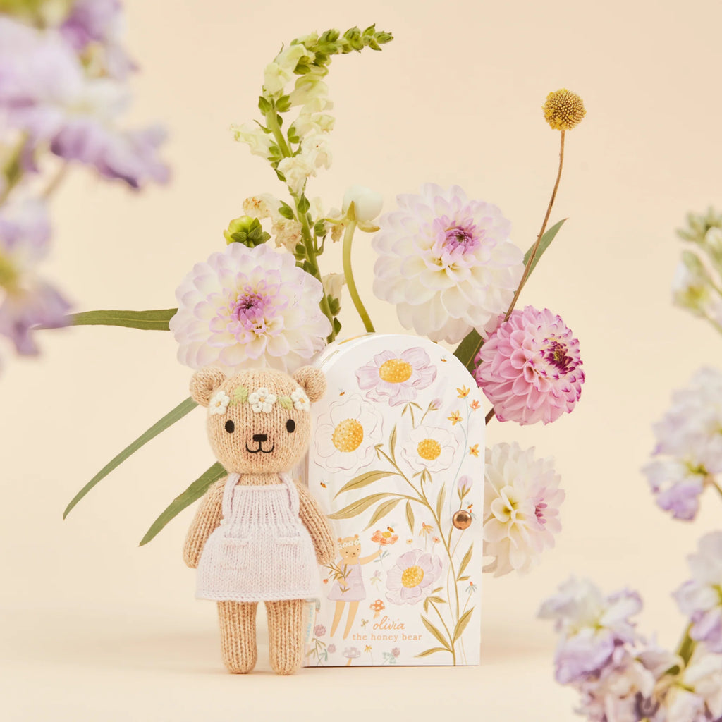 A Cuddle + Kind Tiny Olivia the Honey Bear, in a white dress and hypoallergenic design, sits next to a floral decorative box. In the background, a bouquet of pink and white flowers enhances the light beige setting.