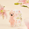 Tiny Penelope the Flamingo, from Cuddle + Kind, is a handmade, safety-certified toy from Peru. It stands before a floral-patterned box with soft-focus pink lilies in the background, enhancing the whimsical scene.