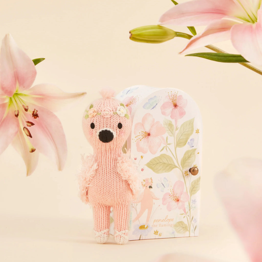 Tiny Penelope the Flamingo, from Cuddle + Kind, is a handmade, safety-certified toy from Peru. It stands before a floral-patterned box with soft-focus pink lilies in the background, enhancing the whimsical scene.
