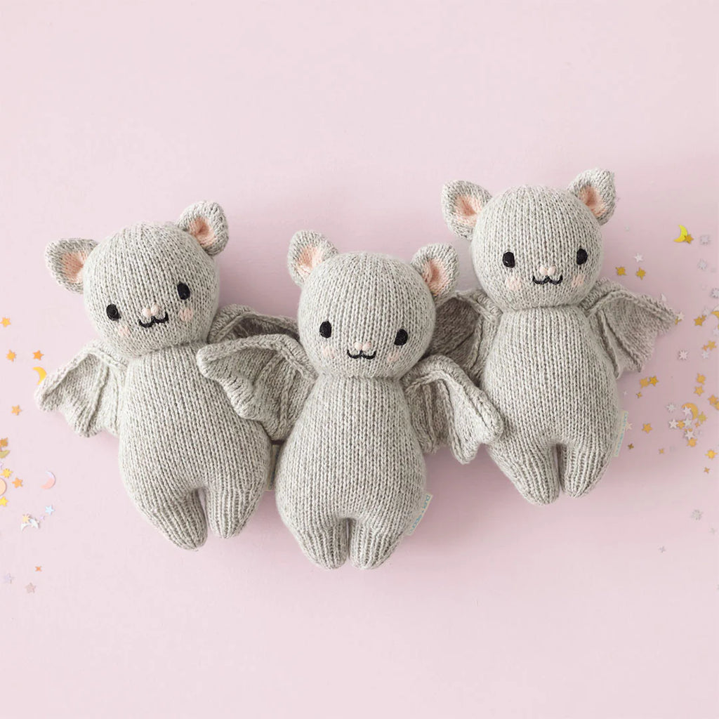 Three Cuddle + Kind Baby Limited Edition Grey Bats with adorable facial expressions and wings outstretched are lined up against a light pink background. Crafted from soft Peruvian cotton yarn, the bats are light gray with pink ear details. Colorful star and moon confetti is scattered around them, making them a delightful collectible.