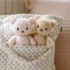 The Cuddle + Kind Baby Bouclé Bear - Honey, crafted with sustainable fair trade practices, sits in a patterned fabric bag next to a soft pink bear, resting against a pale pink textured cushion.