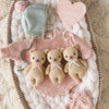 A pink baby romper, mint bonnet, pink booties, and the Cuddle + Kind Baby Honey Bear in Robin’s Egg Blue are arranged on a white blanket beside a heart-shaped "My First Valentine's Day" sign.