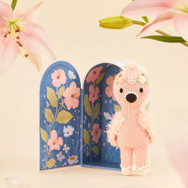 Cuddle + Kind's Tiny Penelope the Flamingo, a handmade pink stuffed animal from Peru, sports a floral headband. It stands in front of an open box with a blue background featuring pink and yellow floral designs, with pink flowers decorating the top corners of this charming scene.