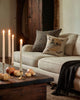 A cozy living room with a beige sofa featuring two decorative pillows, including the Coral & Tusk Supermoon Spell Pillow. A wooden table displays lit candles and small pumpkins, while a dark throw blanket is draped over the sofa's arm.