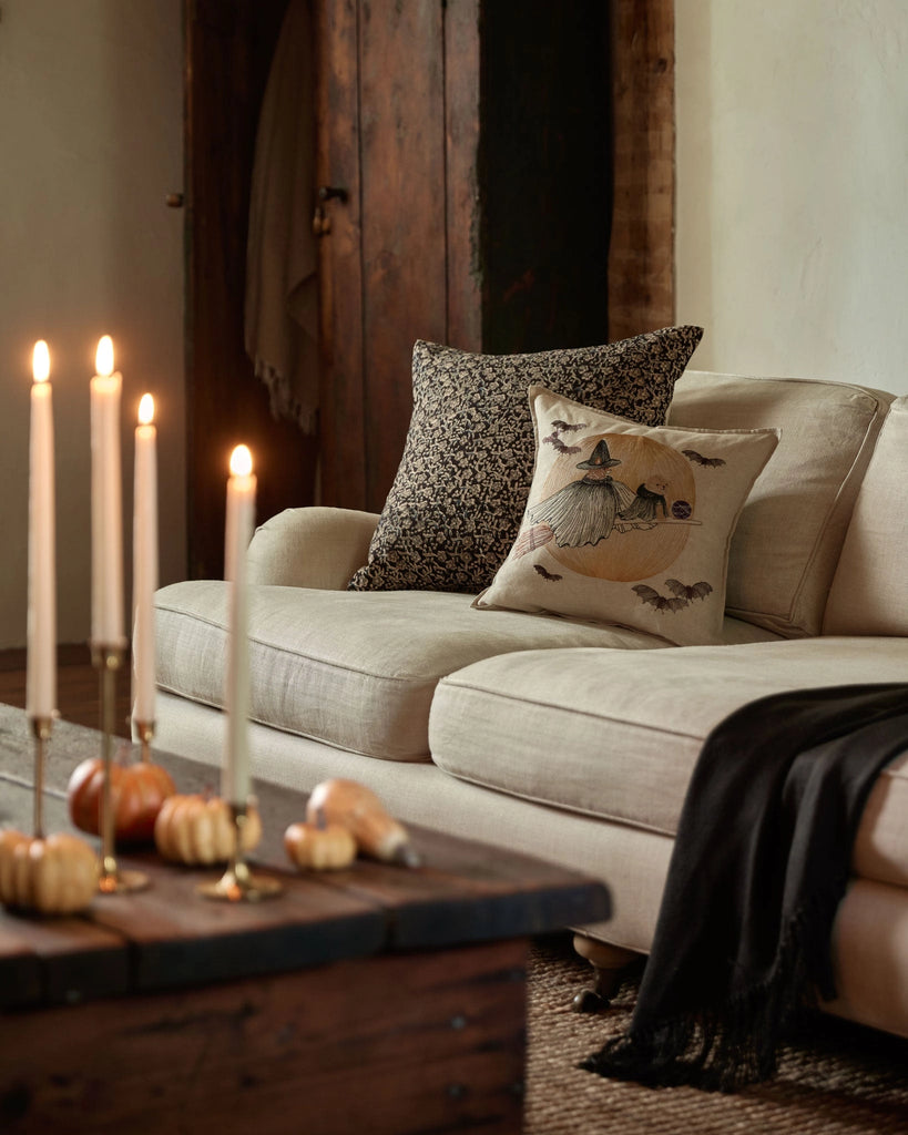 A cozy living room with a beige sofa featuring two decorative pillows, including the Coral & Tusk Supermoon Spell Pillow. A wooden table displays lit candles and small pumpkins, while a dark throw blanket is draped over the sofa's arm.