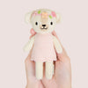 A hand holding an adorable plush toy dog named Tiny Charlotte from Cuddle + Kind, dressed in a pink, hand-knit dress. The dog features embroidered flowers on its head and a smiling face, crafted from non-toxic yarn, set against a soft beige background.