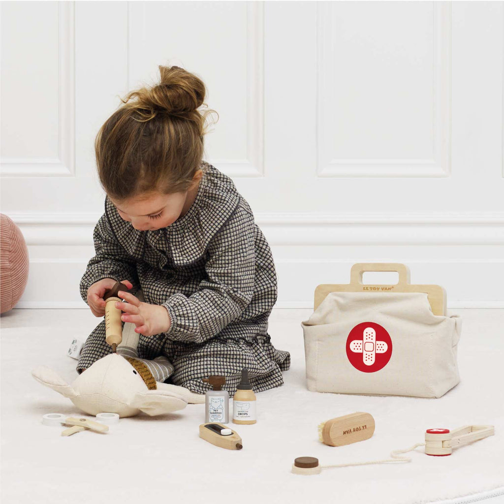 A young child in a checkered dress uses the Pretend Play Animal Vet Set—wooden medical toys and a white doctor’s bag—to care for a plush toy on a light floor in a bright, sunlit room.