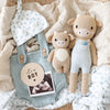 Baby announcement flat lay featuring a hand knit cotton hat, blue overalls, two Cuddle + Kind Asher The Cow plush toys, a round "it's a BOY" sign, and an ultrasound photo arranged on soft neutral bedding.