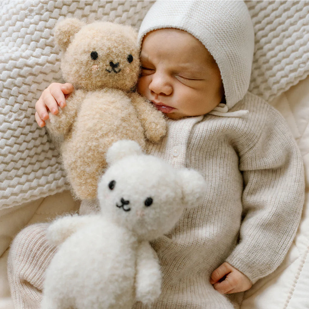 A baby in a soft, cream-colored outfit and knit hat sleeps peacefully holding a beige teddy bear, with the Cuddle + Kind Baby Bouclé Bear - Honey resting nearby on a textured blanket.