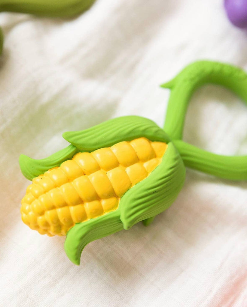 A Corn Rattle Toy Rattle with yellow and green corn design and husk details sits on a white textured fabric surface.
