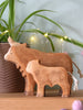 A Handmade Wooden Highland Calf, crafted from sustainable hardwood and finished with plant-based stain, adds cozy charm to a wooden surface with string lights and a potted plant in the background.